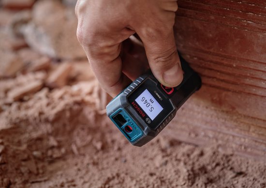 Hand houdt een blauwe Bosch laser-afstandsmeter tegen een ruwe muur, display toont 5,046 m.
