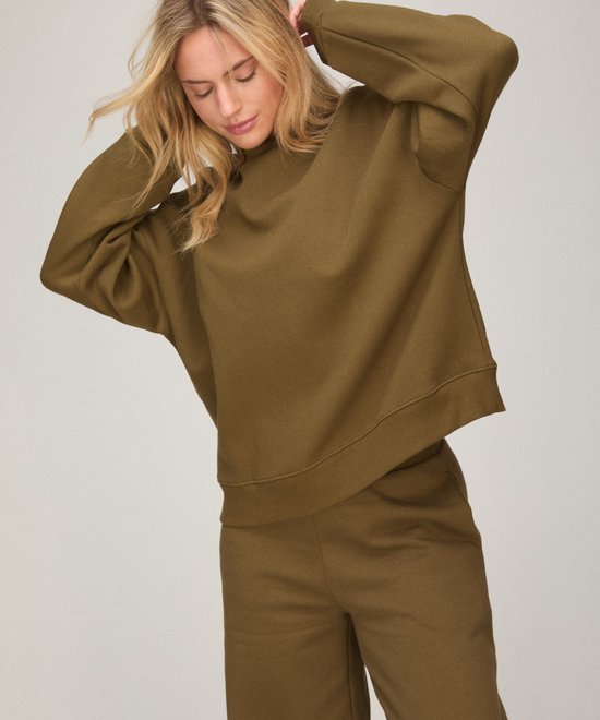 Model draagt een olijfgroene sweatshirt en bijpassende broek, met de handen achter het hoofd.