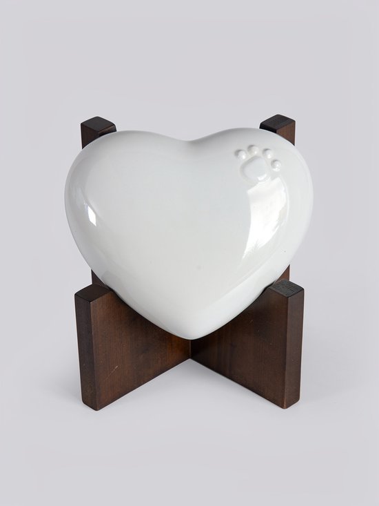 Witte, hartvormige keramische urn met een subtiel dierenpootje, rustend op een donkerhouten standaard.