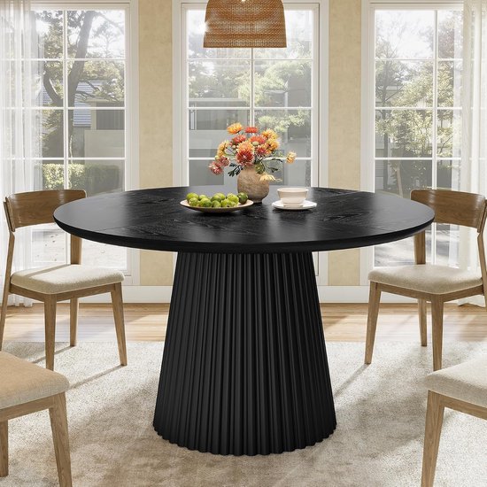 Table à manger ronde noire avec piédestal plissé, décorée d'un vase de fleurs et de fruits, dans une salle à manger lumineuse.