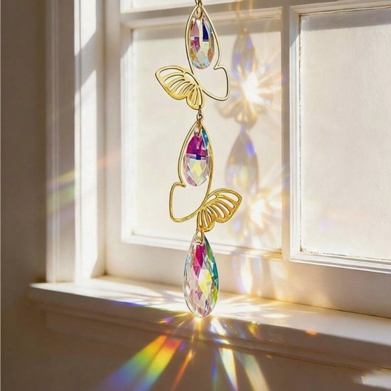 Gouden hanger met vlinders en druppelvormige kristallen, aan een raam, met regenboogeffecten.