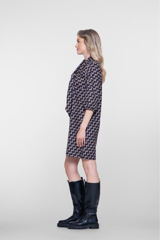Vrouw met blonde haren, profiel, draagt een navy jurk met geometrische print en zwarte laarzen.