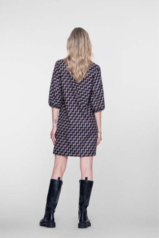 Vrouw met blonde haren, rug naar de camera, draagt een jurk met geometrische print en zwarte laarzen.
