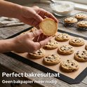 Handen met ringen leggen een ronde cookie op een bakmat vol met chocoladekoekjes.