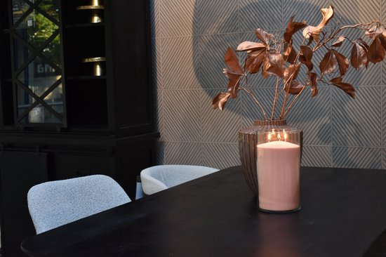 Grote roze kaars in een glazen vaas, naast een koperkleurig boeket op een zwarte tafel.