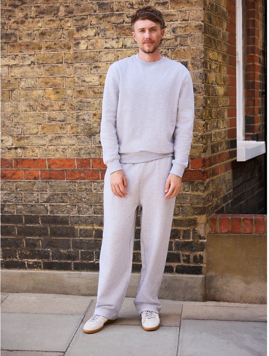 Model draagt een grijs sweatshirt met ronde hals en bijpassende joggingbroek, staand voor een bakstenen muur.