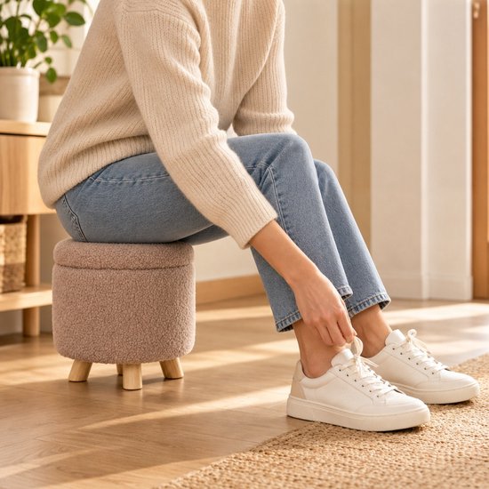 Persoon in beige trui en jeans zit op een bruine teddy poef terwijl ze haar witte sneakers strikt.
