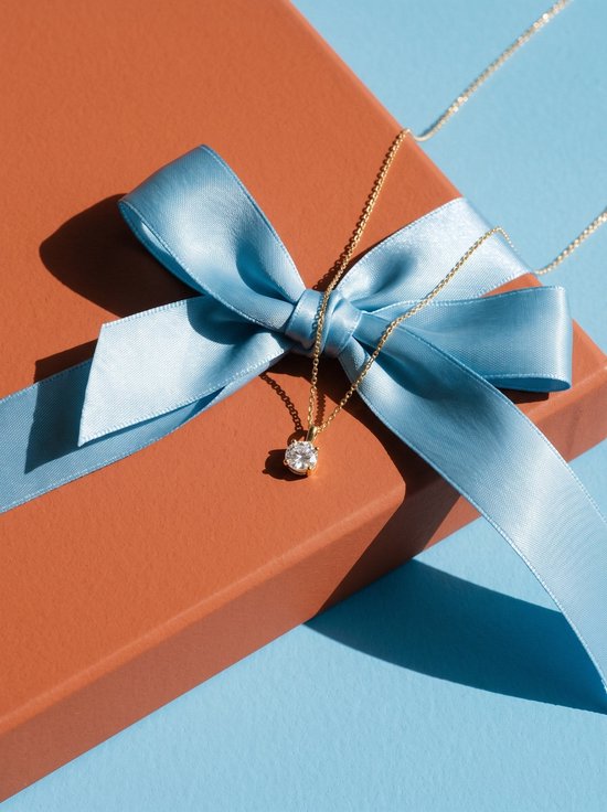 Goudkleurige ketting met ronde zirkonia hanger op een bruine geschenkdoos met een lichtblauwe strik.