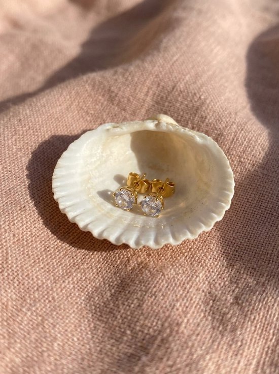 Goudkleurige oorknopjes met zirkonia in een witte schelp op een roze stoffen achtergrond.