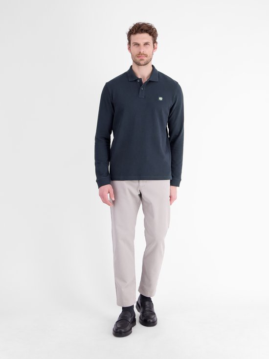 Model draagt een marineblauw poloshirt met lange mouwen, grijsbeige broek en zwarte schoenen.