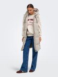 Model staat met handen in de zakken, draagt een lange beige bodywarmer met capuchon over een sweater en jeans.