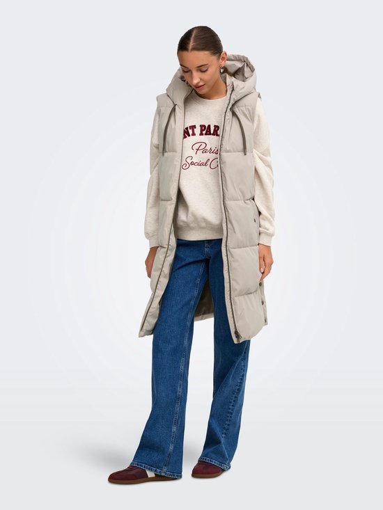Model staat met handen in de zakken, draagt een lange beige bodywarmer met capuchon over een sweater en jeans.