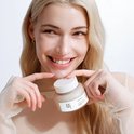 Een blonde vrouw met een glimlach houdt een potje Beauty of Joseon met beide handen vast.