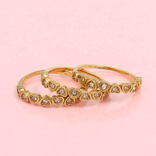Gouden ringen bezet met hartvormige zirkonia stenen op een roze achtergrond.
