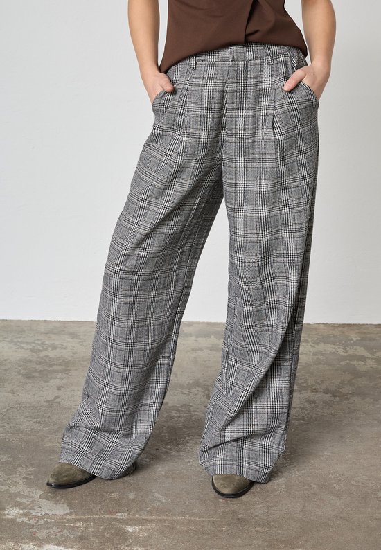Model draagt een grijze pantalon met ruitpatroon, wijde pijpen en bruine top op een betonnen vloer.