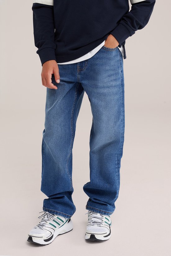 Jongens losse spijkerbroek in blauw denim met witte en groene sneakers.