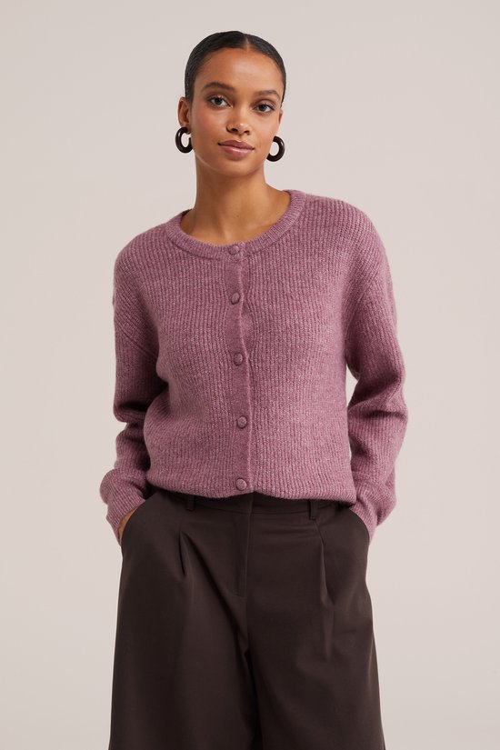 Model draagt een roze, gebreid vest met knoopsluiting en ronde hals, gecombineerd met een donkerbruine broek.