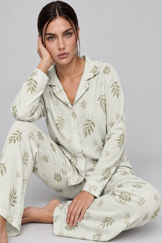 Model zit zittend in een saliegroene pyjamaset met bladerenprint, met de hand bij het gezicht.