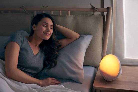 Een vrouw slaapt in een bed bij zacht, geel ochtendlicht van een wekker.