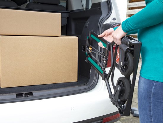 Een persoon laadt een opvouwbare steekwagen met groene handgrepen in de kofferbak van een auto, naast twee dozen.