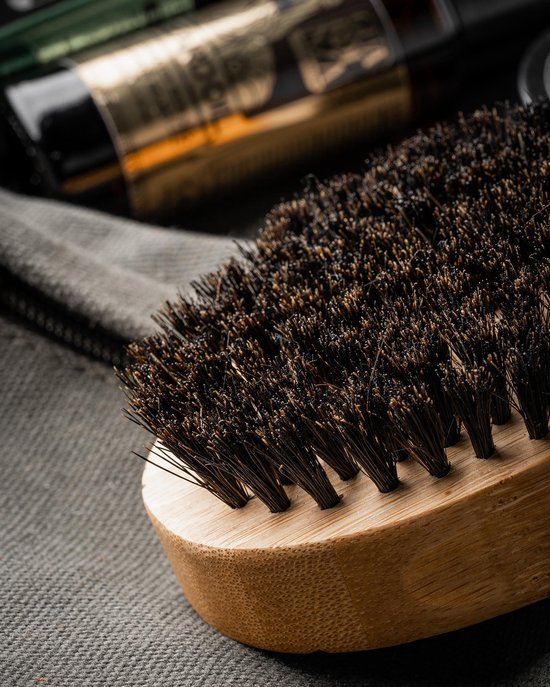 Close-up van een ovale bamboe baardborstel met dichte, donkere haren op een textiel achtergrond.
