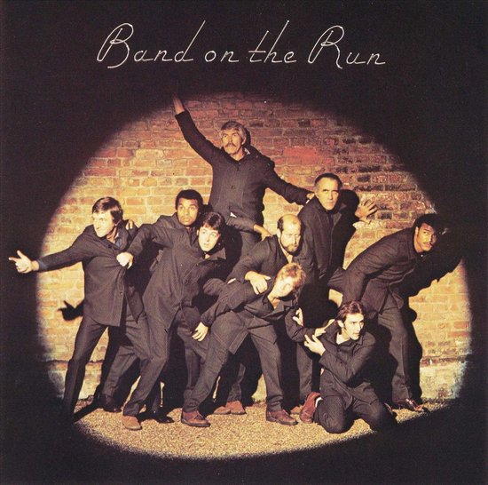 Albumhoes 'Band on the Run' met negen mannen in donkere pakken in een spotlight voor een bakstenen muur.