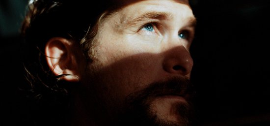 Close-up van een man met blauwe ogen, een baard en snor. Een schaduw valt over zijn gezicht.