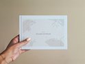 Een hand die een gesloten kraamvisiteboek vasthoudt. De omslag is licht beige met abstracte vormen en botanische illustraties.