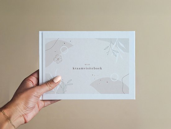 Een hand die een gesloten kraamvisiteboek vasthoudt. De omslag is licht beige met abstracte vormen en botanische illustraties.