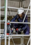 Vrouw in blauw werkpak en helm, op steiger, houdt CEE-stekker vast bij kabelhaspel.