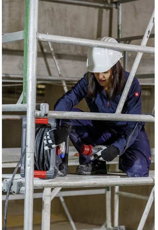 Vrouw in blauw werkpak en helm, op steiger, houdt CEE-stekker vast bij kabelhaspel.