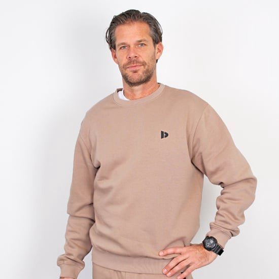 Man in een donkerbeige sweater met zwart logo op de borst.