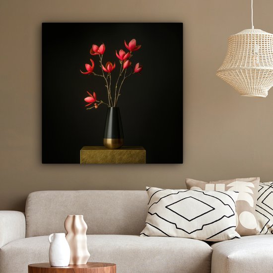 Rode bloemen in een zwarte vaas op een gouden sokkel, in een woonkamer met een bank en kussens.