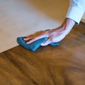 Une main avec un chiffon en microfibre bleu-gris nettoie un plateau de table en bois.