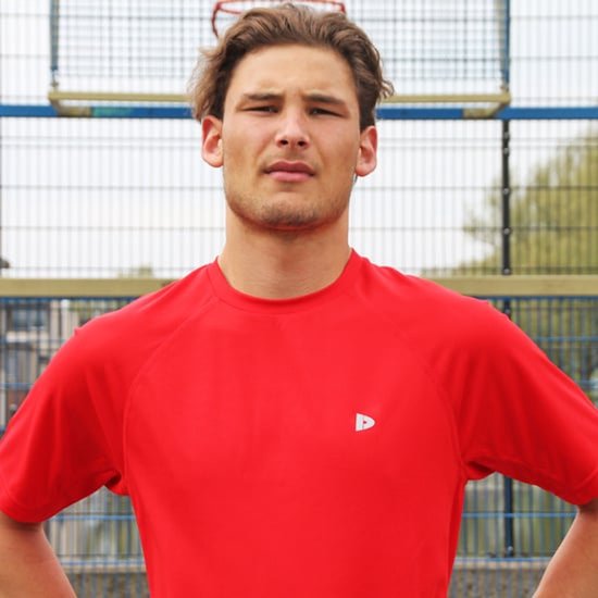 Man draagt een rood sportshirt met korte mouwen, met de handen in de zij.