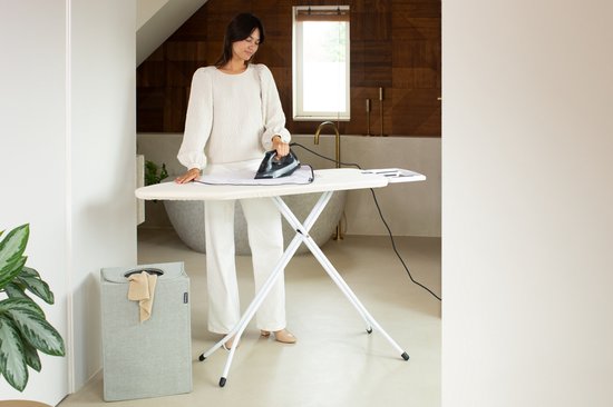 Vrouw strijkt met een strijkijzer op een crèmekleurige strijkplank in een badkamer.