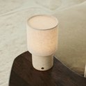 Tafellamp met beige linnenkap en cilindrische travertijnen voet op een donker tafelblad.