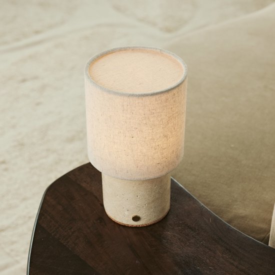 Tafellamp met beige linnenkap en cilindrische travertijnen voet op een donker tafelblad.