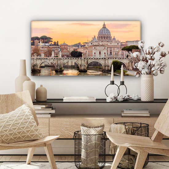 Canvas van de Sint-Pietersbasiliek en de Ponte Sant'Angelo in Rome bij zonsondergang, gepresenteerd in een woonkamer met houten meubels.