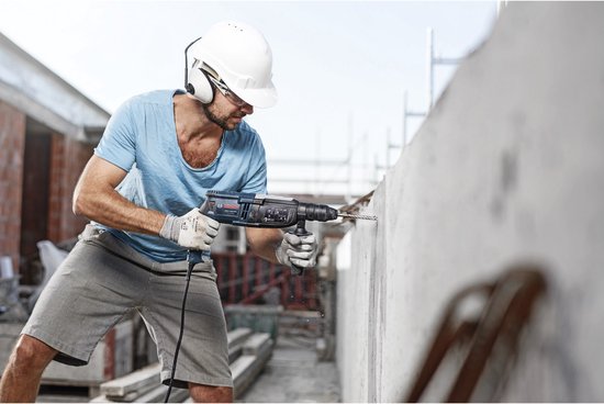 Een man met helm boort met een blauwe Bosch boor in een betonnen muur.