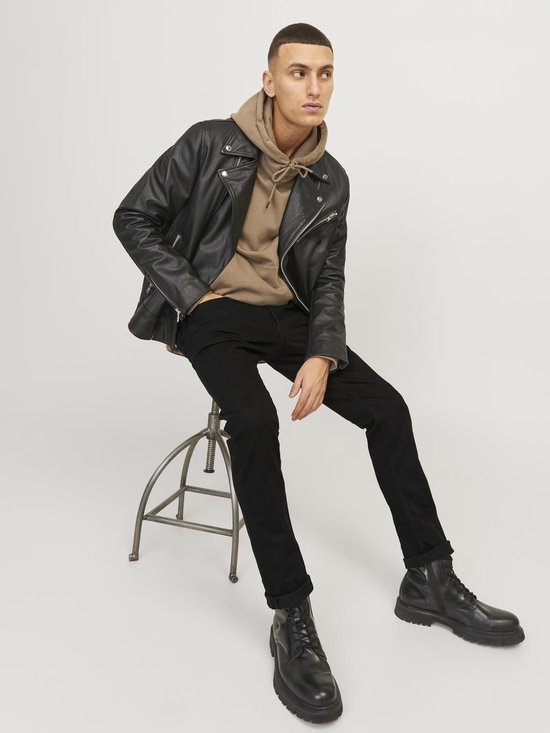Man in zwarte jeans, bruine hoodie en zwart leren jack, zittend op een krukje.