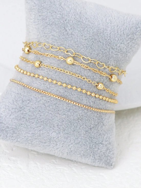 Vijf gouden armbandjes met diverse schakels, waaronder hartjes en kraaltjes, gestapeld op een grijs kussen.