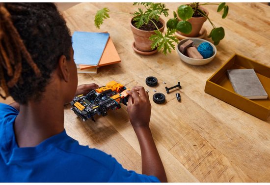 Kind bouwt een oranje en zwarte LEGO Technic NEOM McLaren Extreme E Team auto op een houten tafel.