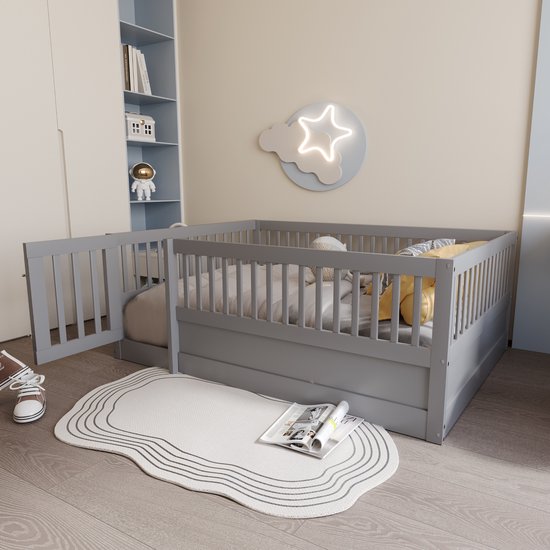 Grijs kinderbedje met hekken en een deurtje in een kinderkamer met een wit vloerkleed en een kast.
