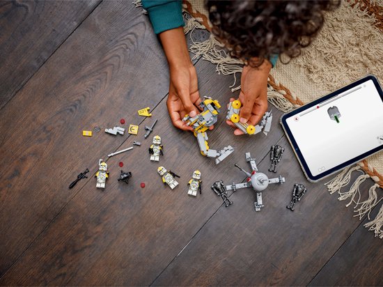 Mains d'enfant construisant un AT-RT walker LEGO Star Wars avec des pièces jaunes et grises, à côté de petites figurines LEGO et d'une tablette.