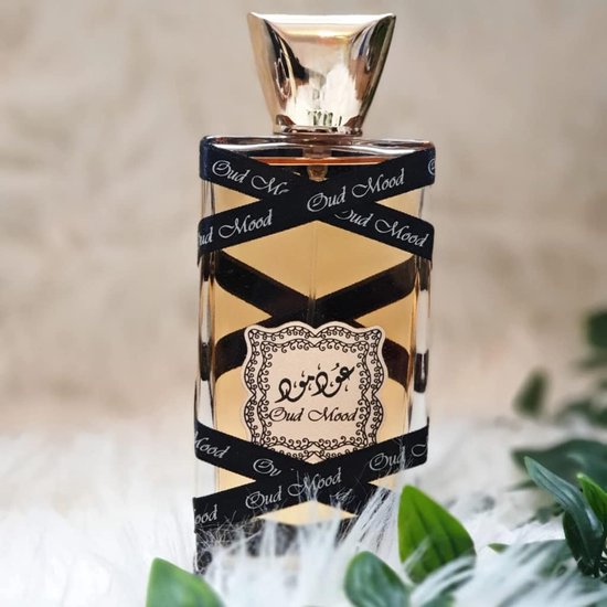 Oud Mood parfumfles met gouden dop, omwikkeld met zwart lint met "Oud Mood" erop. Op een witte ondergrond met groene bladeren.