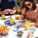 Des gens jouent au domino et aux cartes à une table avec des fruits et des boissons, dont deux verres de boisson jaune.