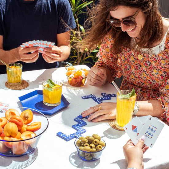 Des gens jouent au domino et aux cartes à une table avec des fruits et des boissons, dont deux verres de boisson jaune.