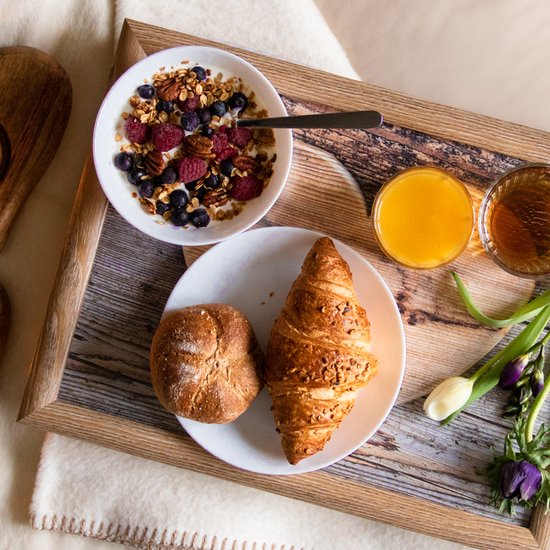 Ontbijt op een houten dienblad: croissant, broodje, yoghurt met fruit, jus d'orange en thee. Met bloemen.