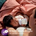 Een baby drinkt uit een Philips Avent fles, liggend op een roze deken. De tekst luidt 'El bebe adapta el ritmo de alimentación'.